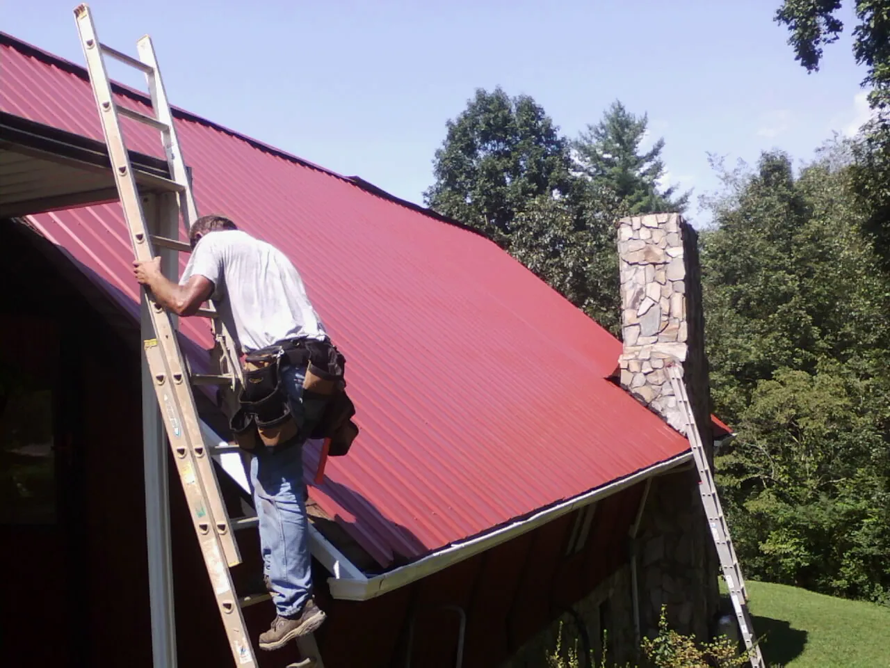 Roofer on a ladder beside a red metal roof and stone chimney — Soffit & Fascia Repair by Roofing Brothers in Cold Spring